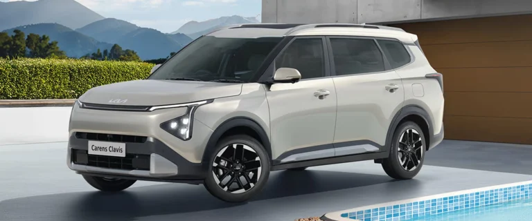 A pristine white Kia Carens SUV parked outdoors with mountains and lush greenery in the background.