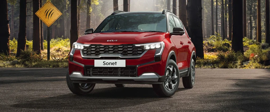 A vibrant red Kia Sonet SUV with a black roof parked on a forest road, with tall trees and a yellow warning sign in the background.